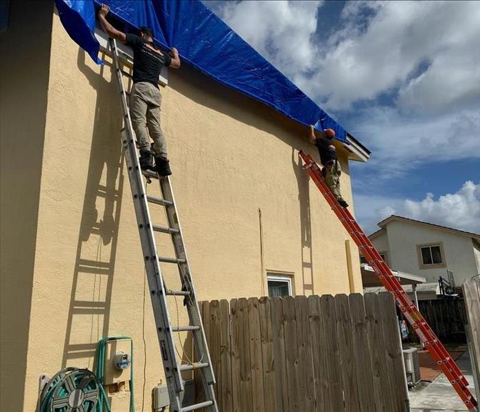 Two men on separate ladders holding on to a blue tarp. 
